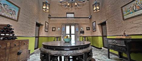 A room with a round wooden table in the center, a wooden cabinet with wine bottles on the left, and a green wall with framed pictures.