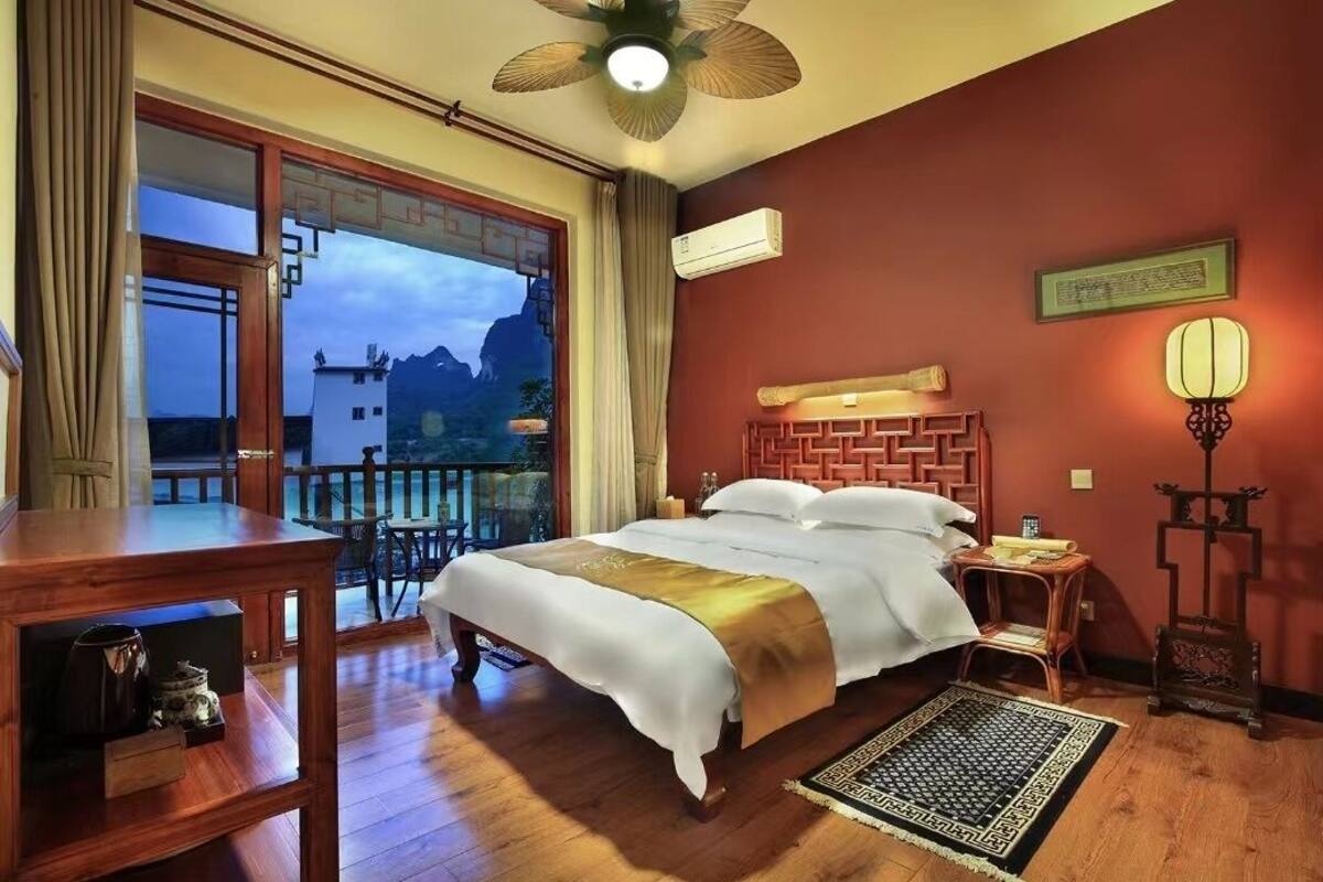 A hotel room with a bed, wooden furniture, a balcony with a view of mountains, and a traditional-style lamp.