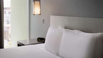 A hotel room with a large bed, white linens, a nightstand with a digital clock, and a wall-mounted lamp.