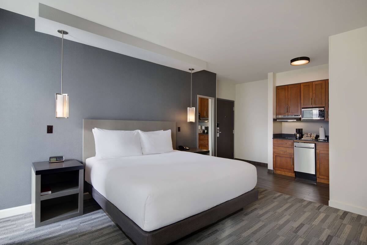 Une chambre d’hôtel moderne dotée d’un grand lit, d’une table de chevet, d’une petite table d’appoint, d’un bureau intégré, d’un four à micro-ondes et d’une petite kitchenette.