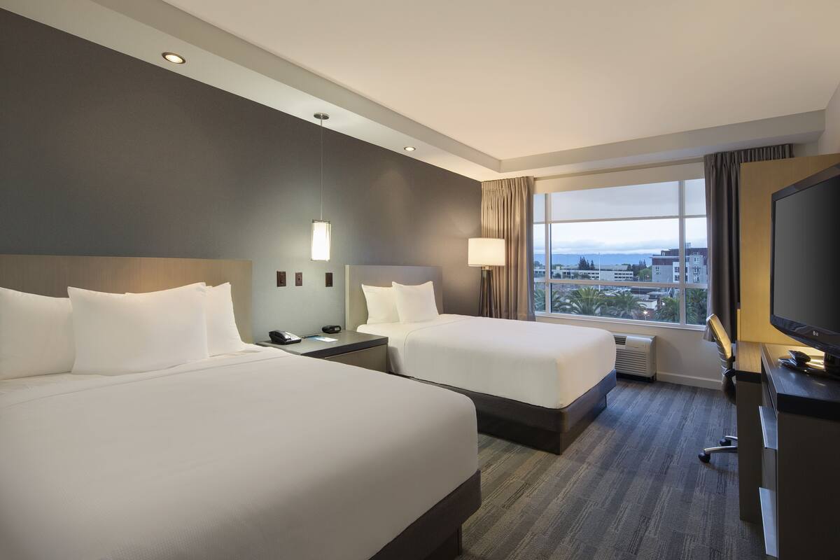 Une chambre d’hôtel avec deux lits, une télévision, un bureau et une grande fenêtre donnant sur la ville.