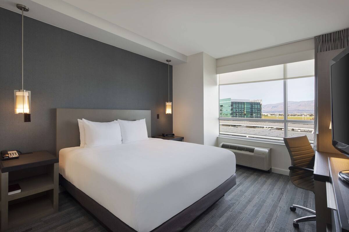 Une chambre d’hôtel dotée d’un grand lit, de tables de chevet, d’un bureau avec une chaise et d’une grande fenêtre donnant sur la ville.