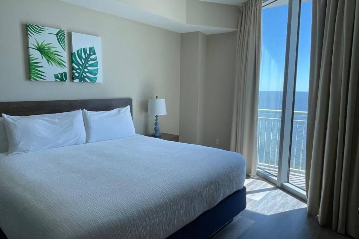 A hotel room with a large bed, a nightstand with a lamp, and a view of the ocean through a balcony.