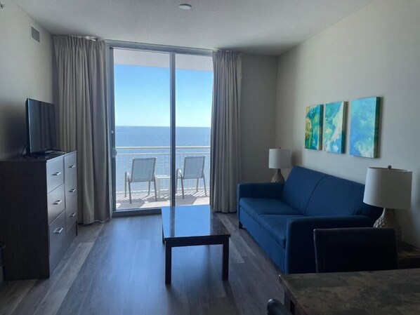 A hotel room with a balcony, a blue sofa, a small table, and a TV on a dresser.