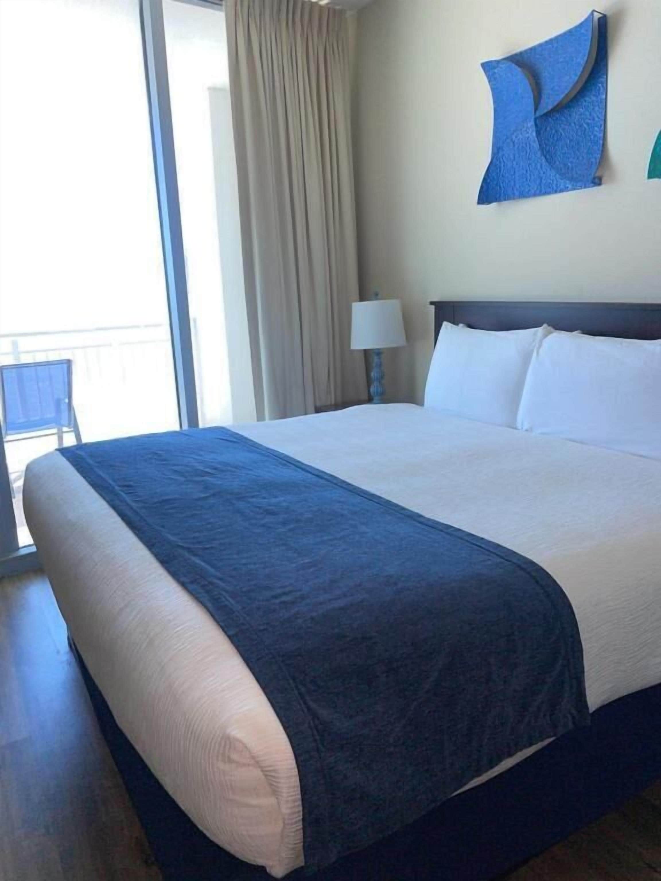 Habitación de hotel con una cama grande, una mesita de noche con lámpara, vistas al exterior y un adorno azul en la pared.