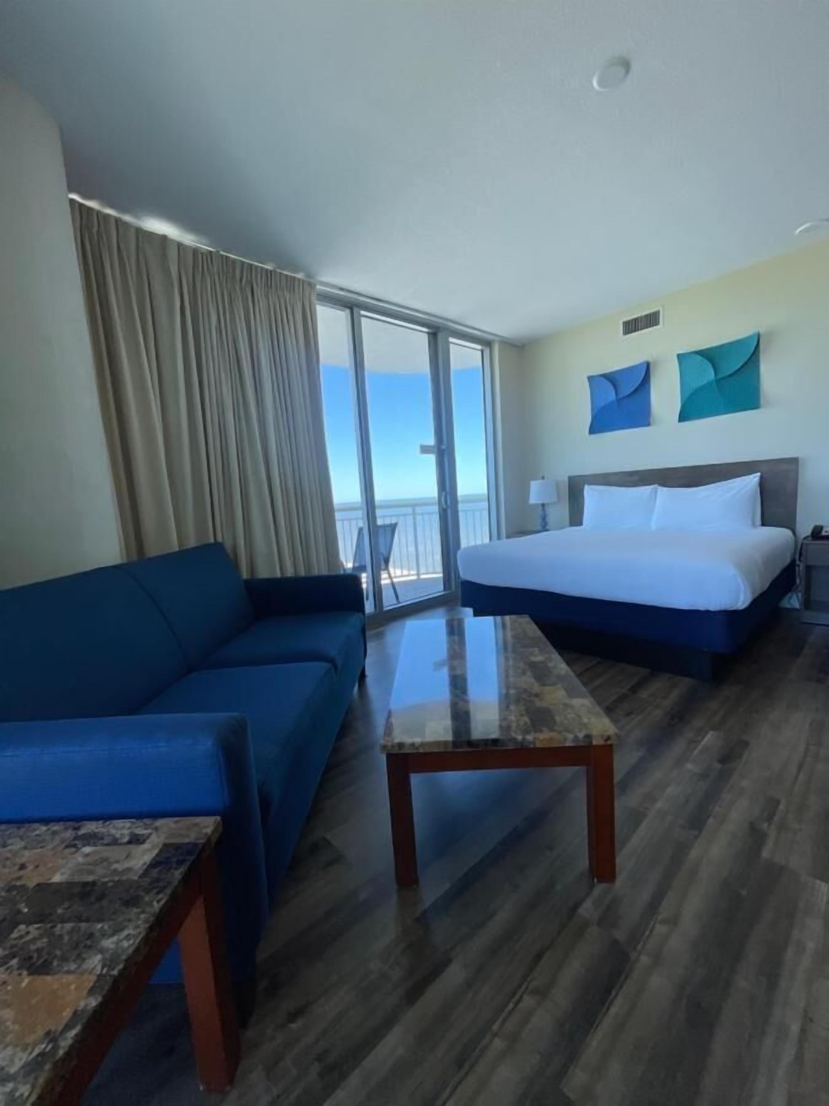 A hotel room with a bed, a blue sofa, a glass coffee table, and a view of the ocean through large windows.