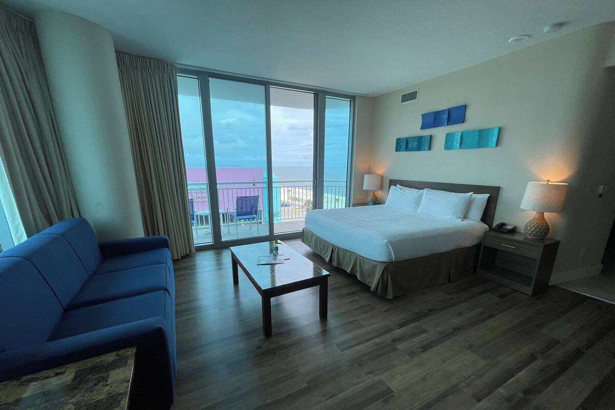 A hotel room with a large bed, a blue armchair, a small table, and a nightstand with a lamp. There is a balcony with a view of the city.