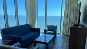 Una habitación con un sofá azul, una mesita, un televisor sobre un mueble y vista al océano a través de amplias ventanas.