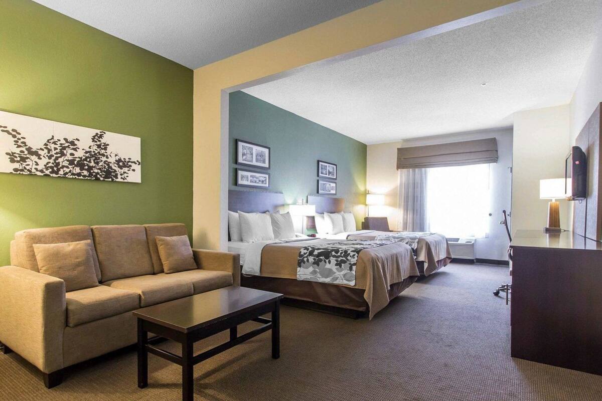 A hotel room with a sofa, two beds, a desk, and a large window.