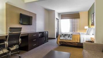 A hotel room with a bed, a desk with a chair, a television mounted on the wall, and a window with curtains.