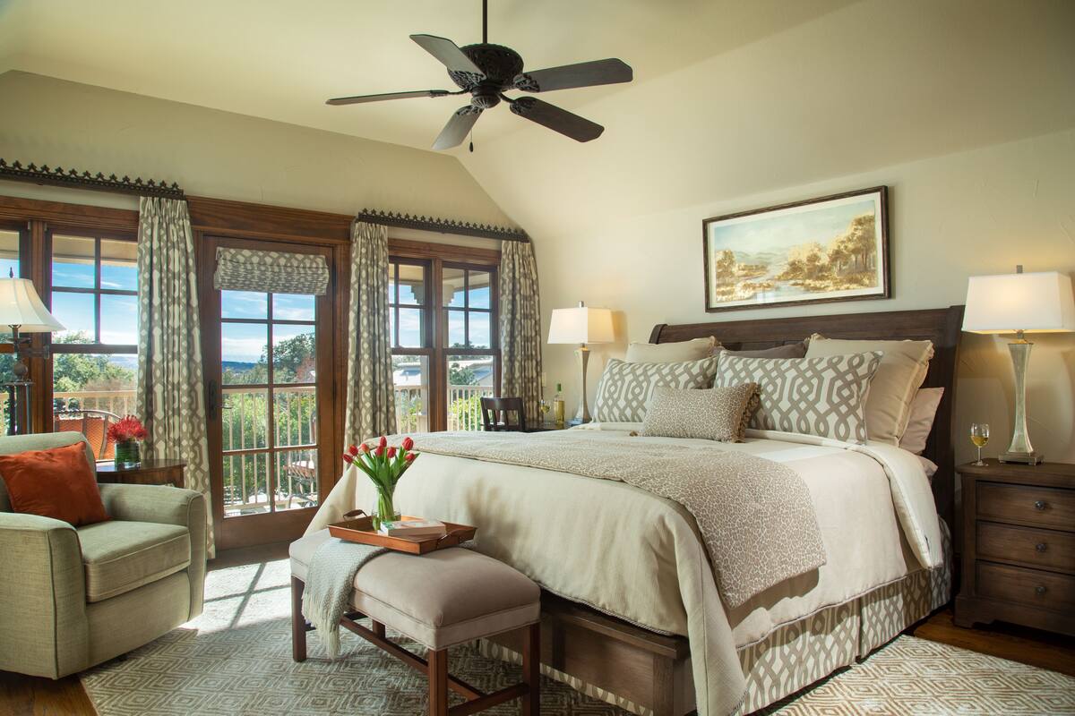 A bedroom with a large bed, a ceiling fan, a nightstand, a chair, and a window with curtains.