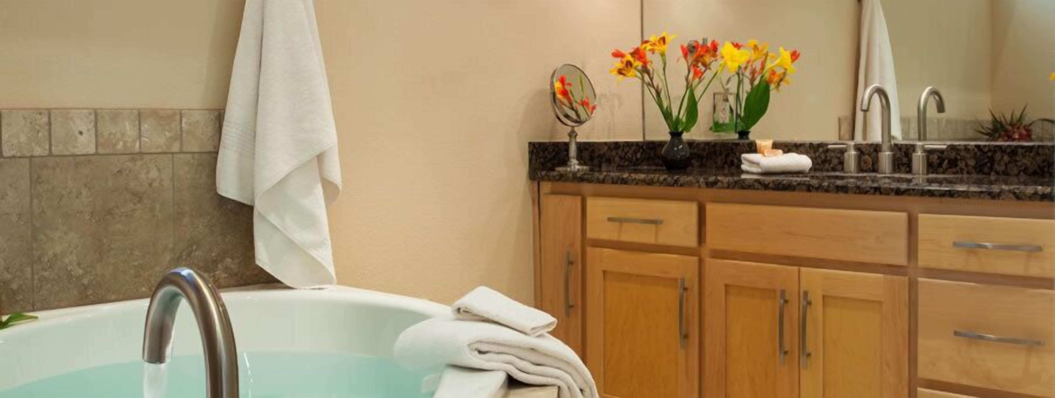 Un baño moderno con bañera, muebles de madera, encimera de granito, espejo y un florero con flores.