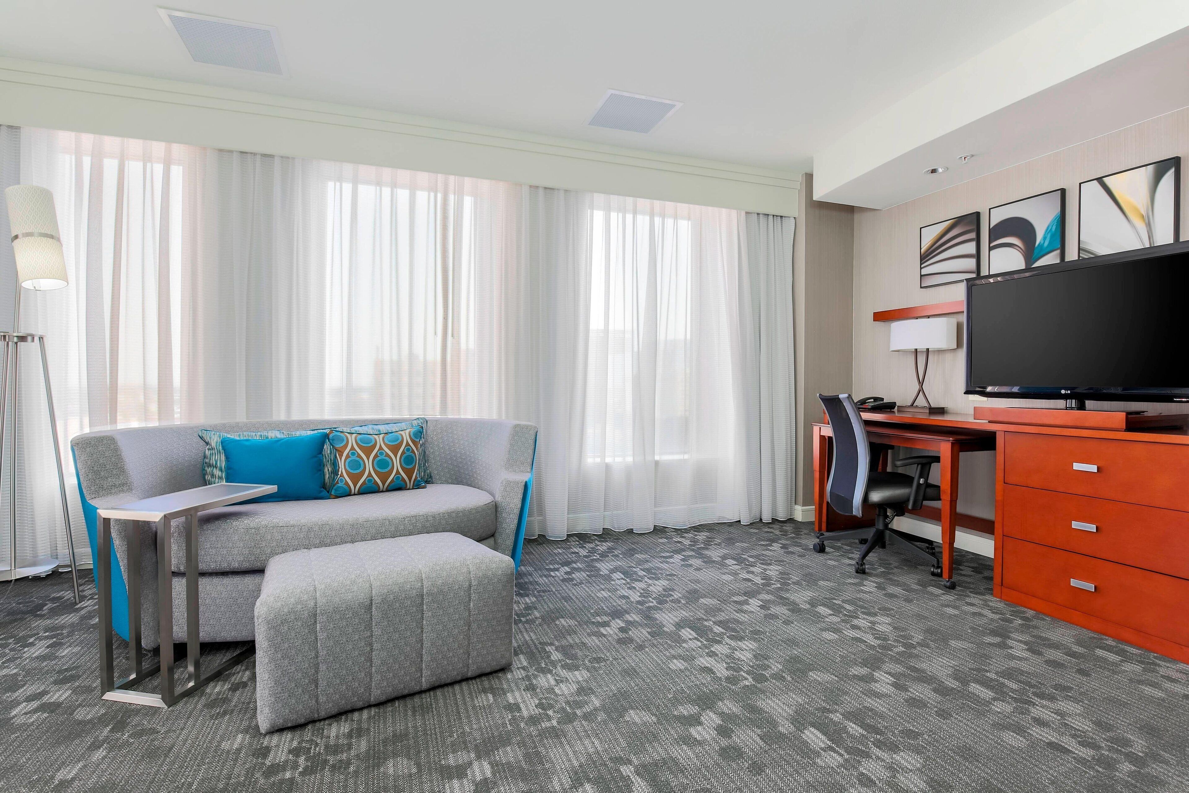 A hotel room with a grey sofa, a small table, a desk with a chair, a TV, and a large window with sheer curtains.