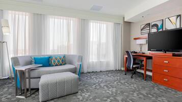 A hotel room with a grey sofa, a small table, a desk with a chair, a TV, and a large window with sheer curtains.
