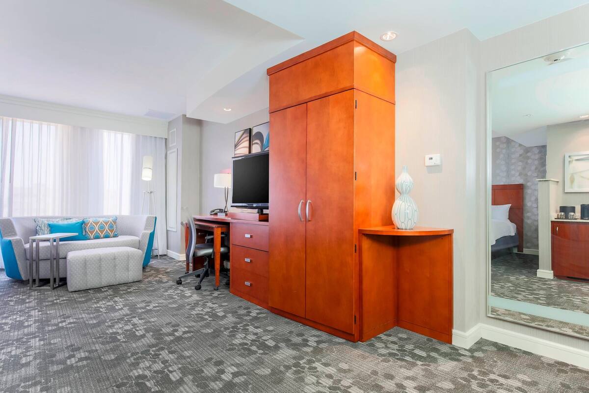 A hotel room with a grey sofa, a wooden wardrobe, a desk with a computer, and a large mirror.