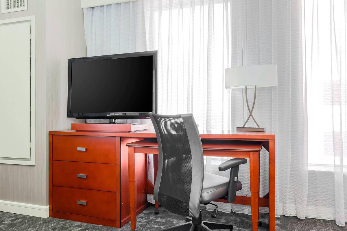 A hotel room with a wooden desk, a television, a chair, and a lamp.