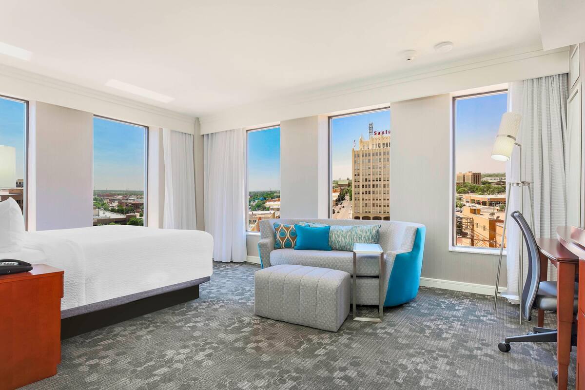 A hotel room with a large bed, a sofa, an ottoman, a desk, and a chair. There are large windows with a city view.