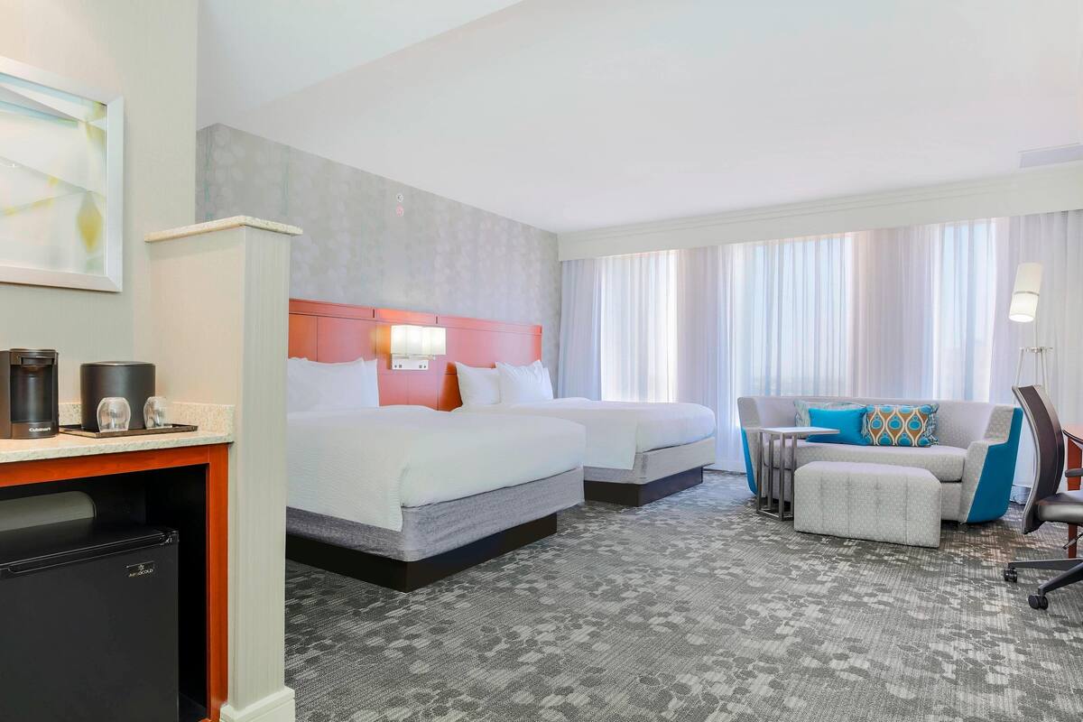 A hotel room with two beds, a desk with a coffee maker, a mini-fridge, and a seating area with a couch and ottoman.