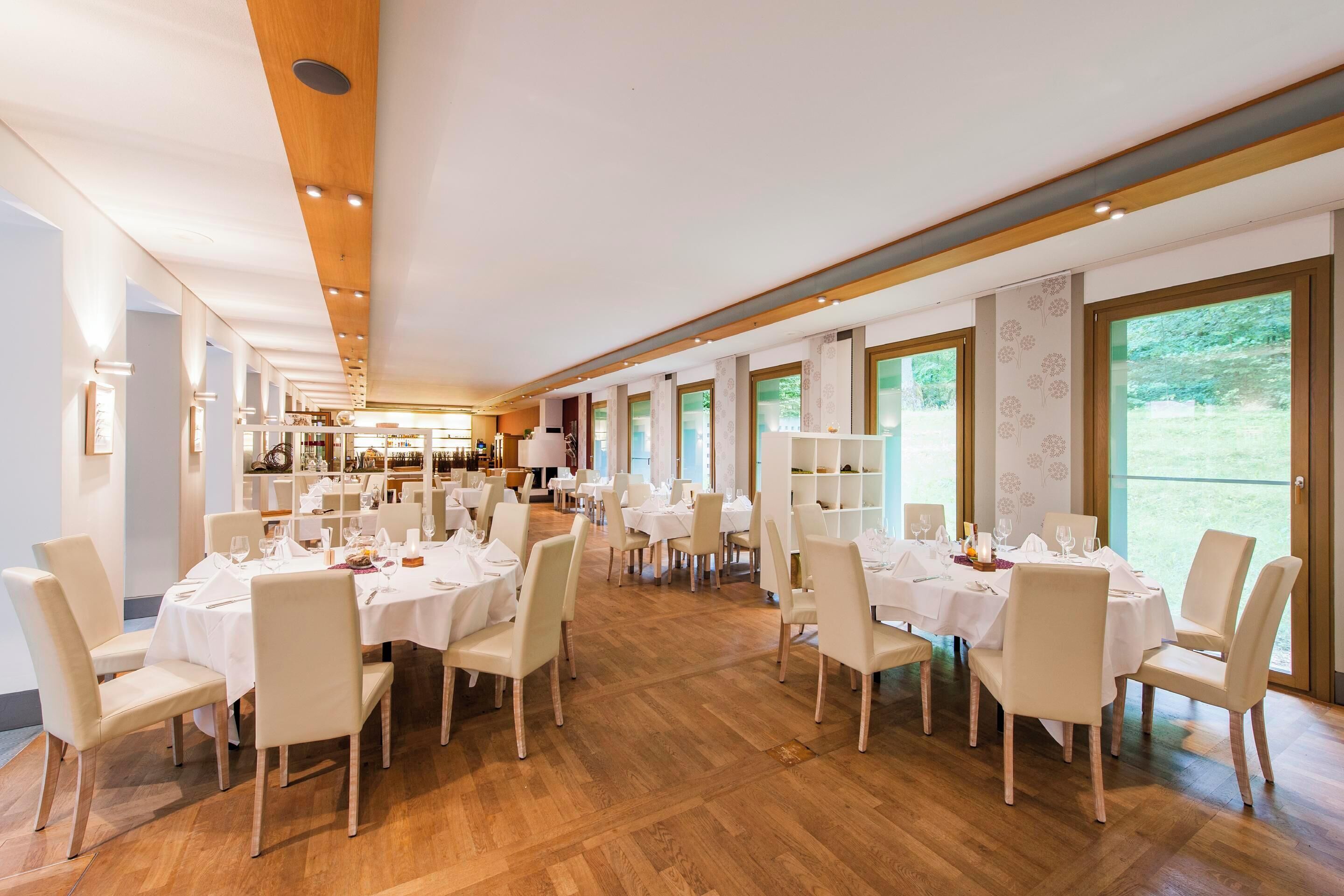 A spacious restaurant with multiple tables and chairs, large windows, and a wooden ceiling.