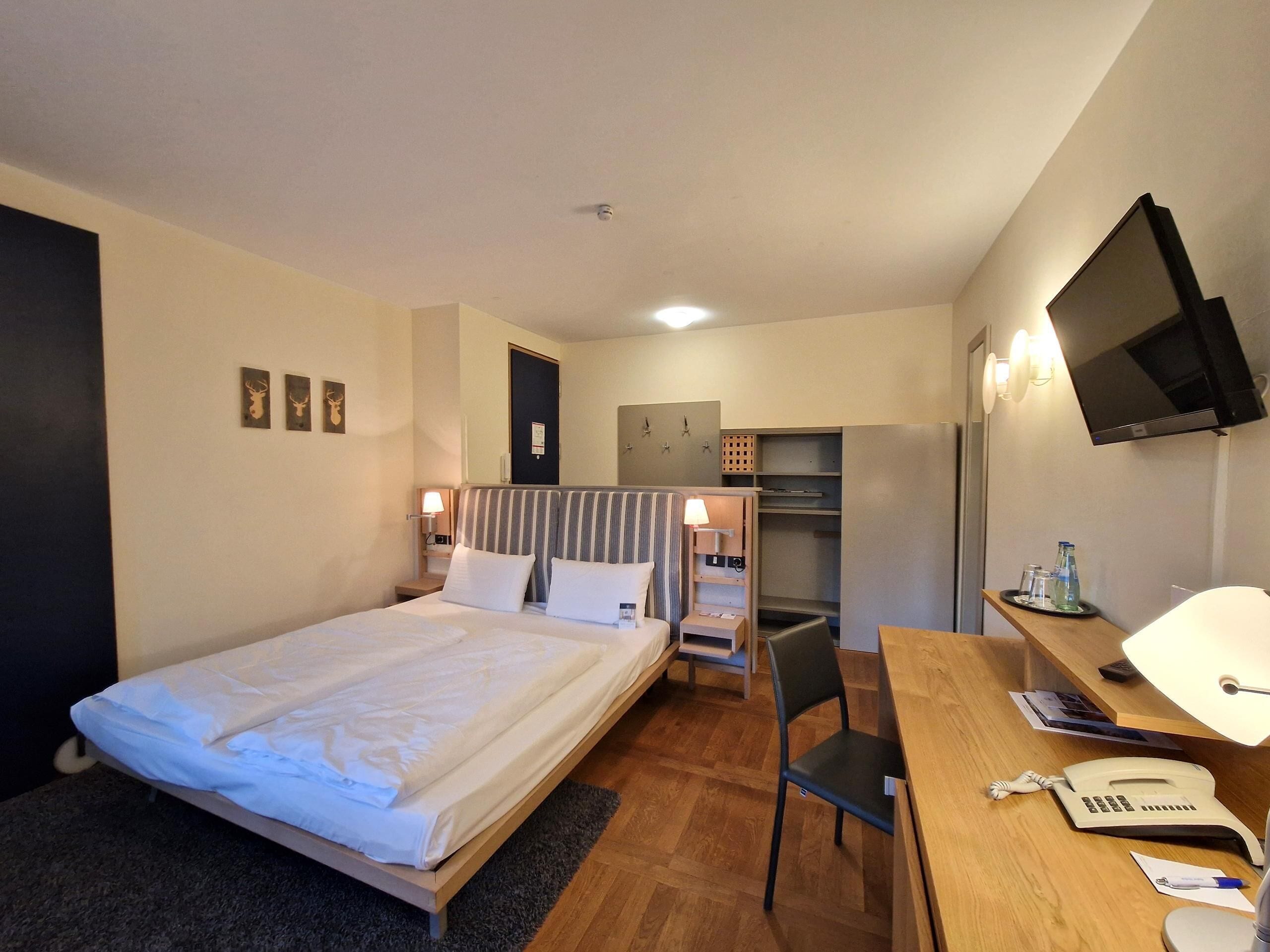 Ein Hotelzimmer mit Bett, Schreibtisch, Stuhl, Fernseher und einer kleinen Küchenzeile.