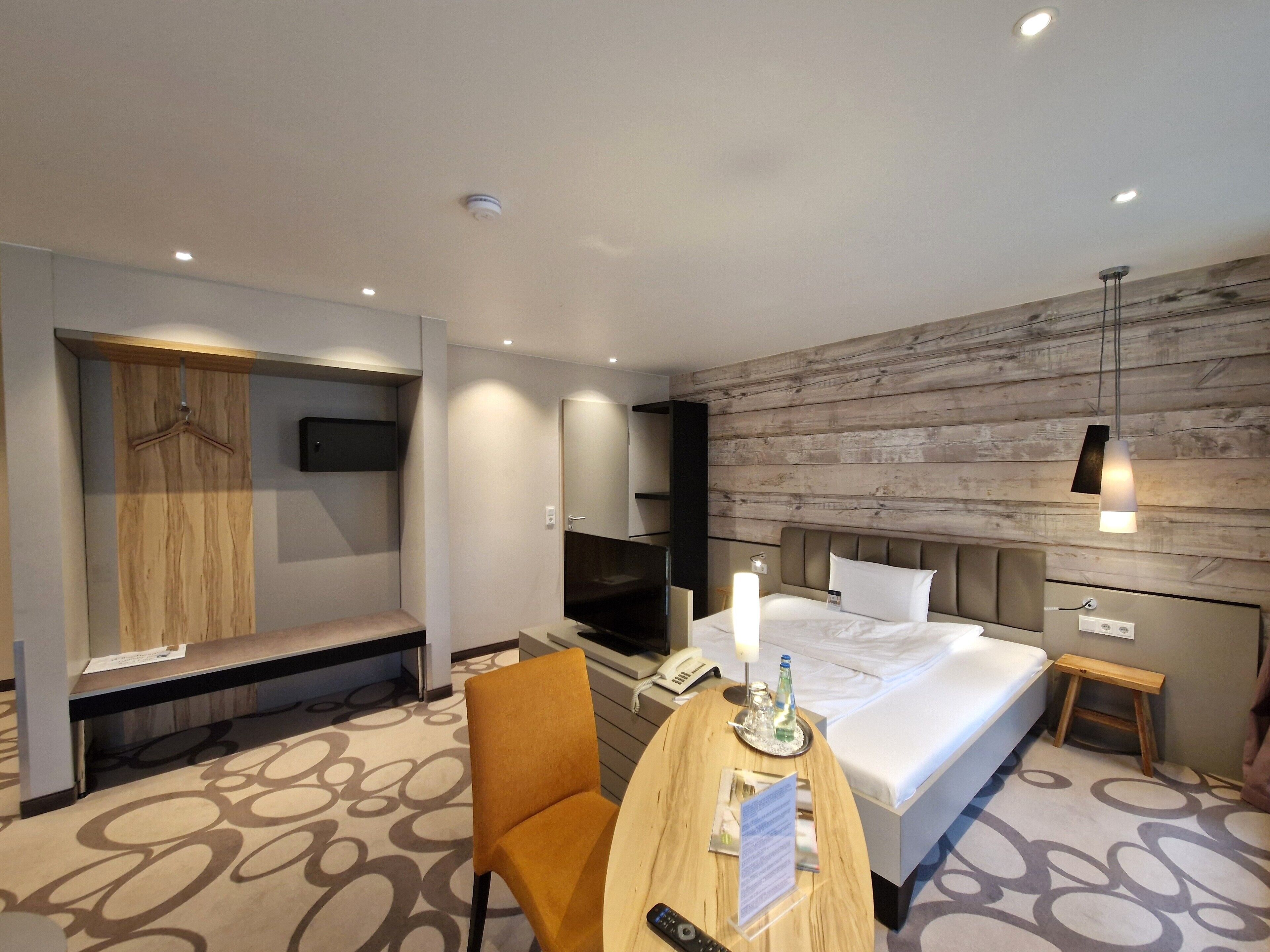 A modern hotel room with a bed, a desk with a chair, a TV, a wooden wardrobe, and a wooden wall panel.