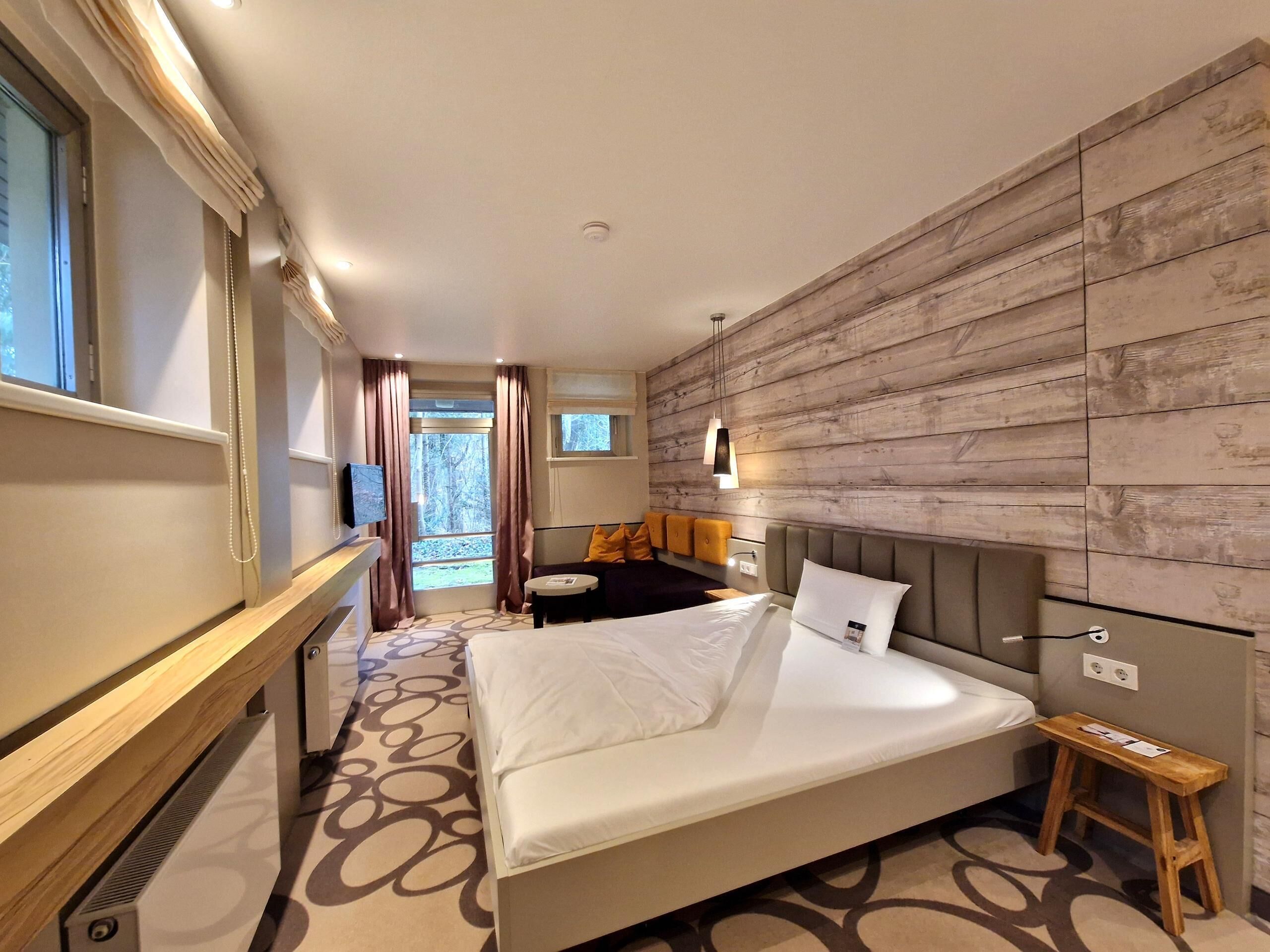 Quarto de hotel moderno com uma cama grande, painel de parede em madeira, um pequeno espaço de estar e uma janela com cortinas.