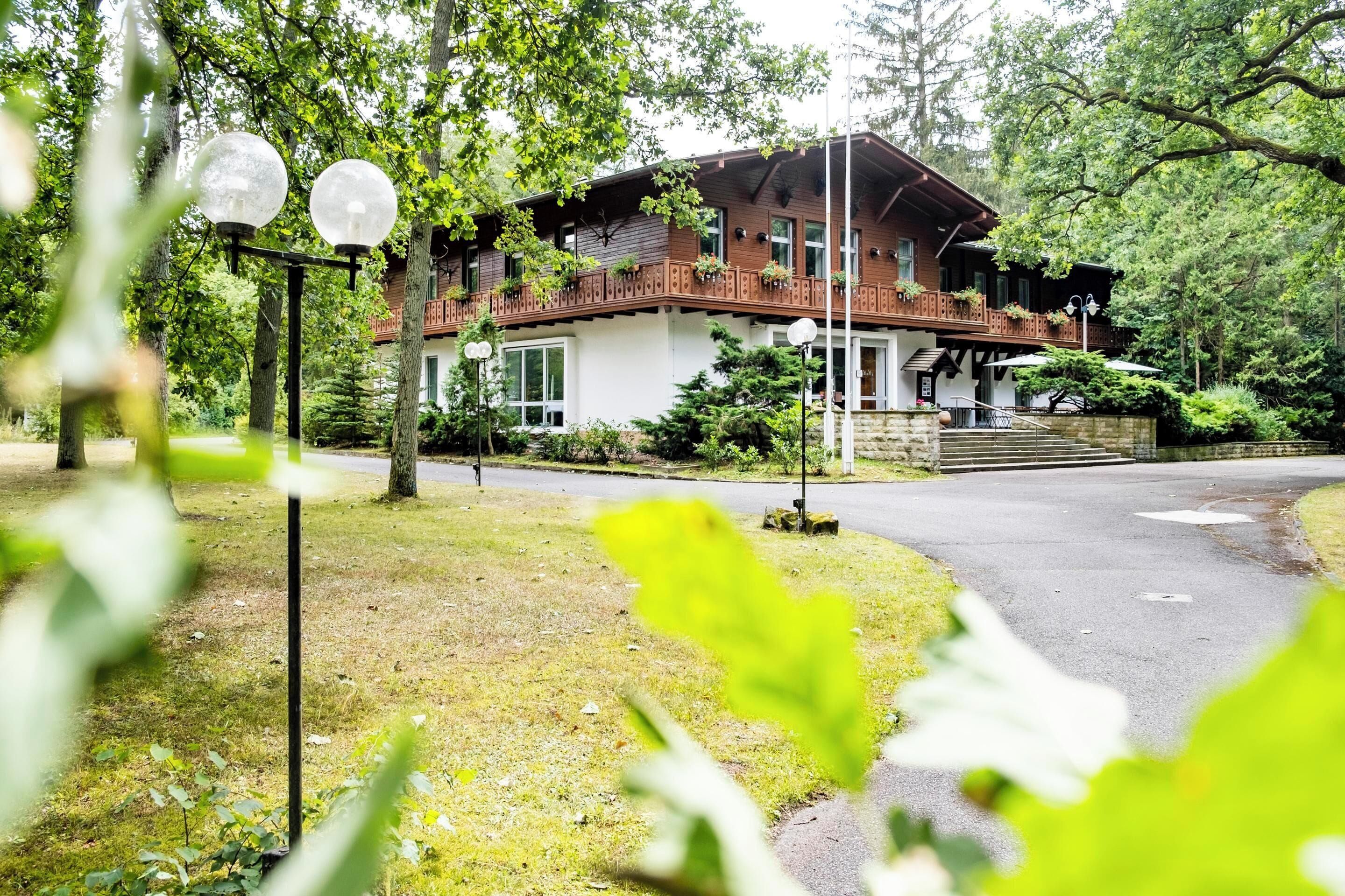 A two-story wooden house with a balcony, surrounded by trees and a paved driveway.