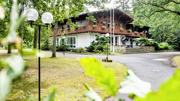 Een houten huis met twee verdiepingen en een balkon, omgeven door bomen en een verharde oprit.