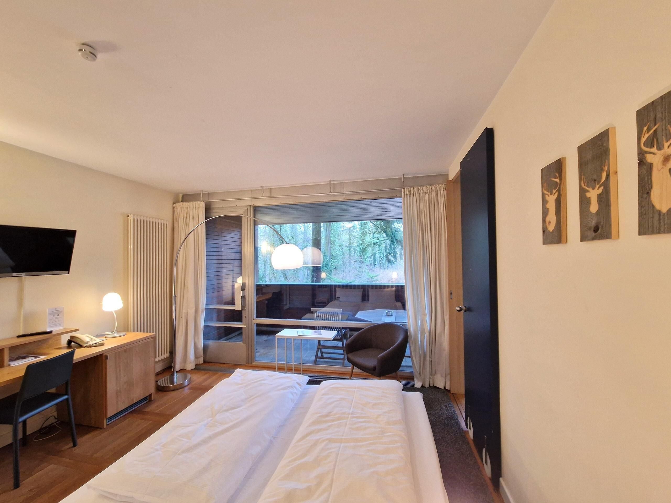 A hotel room with a bed, desk, chair, and a TV. There is a window with curtains and a view of trees.