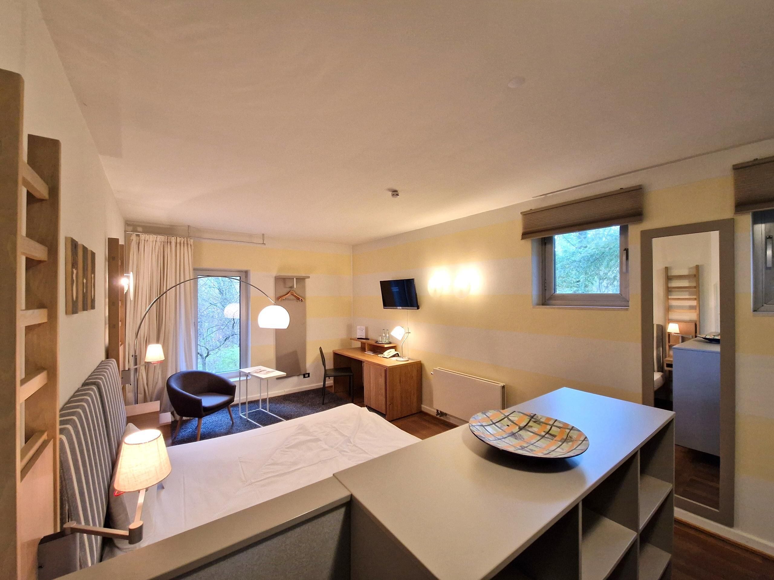 Habitación de hotel con cama, escritorio con computadora, silla, televisor y ventana con cortinas.