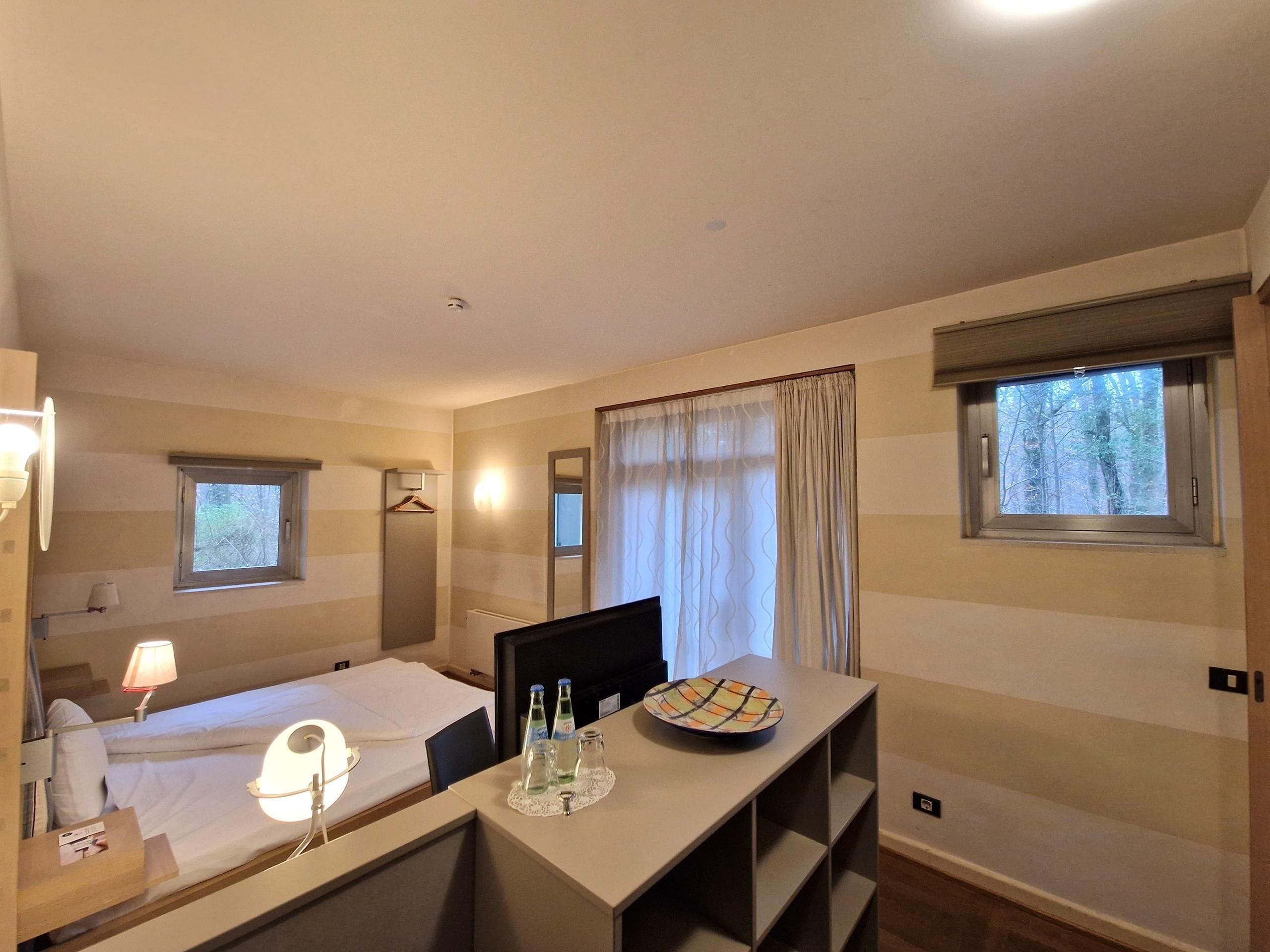 A hotel room with a bed, a desk with a plate and bottles, a mirror, and two windows.