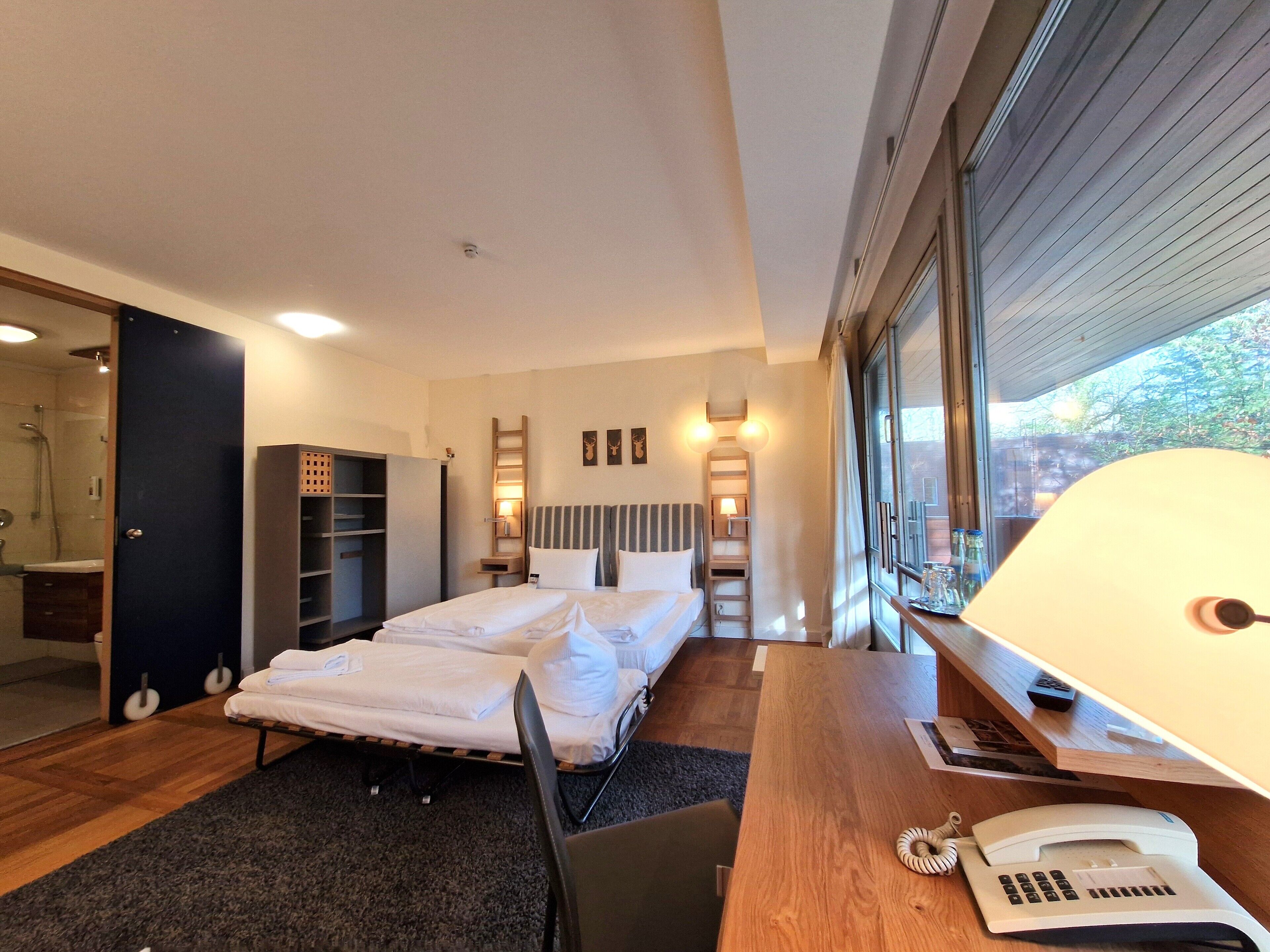 Habitación de hotel moderna con cama, escritorio con lámpara, teléfono y vistas al exterior.