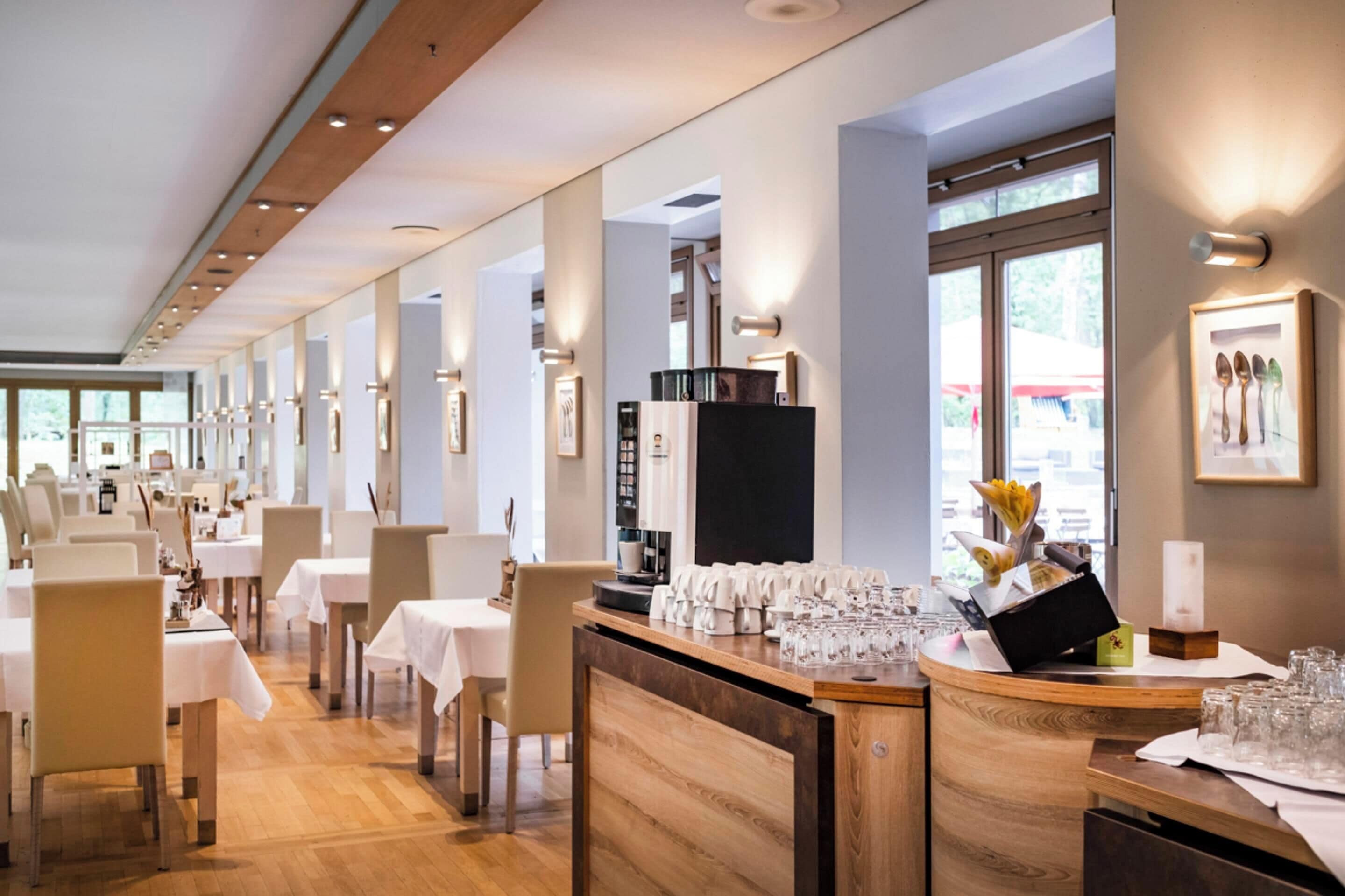 Un restaurante moderno con suelo de madera, mesas y sillas blancas. Hay cuadros en las paredes y una barra con vasos y utensilios.