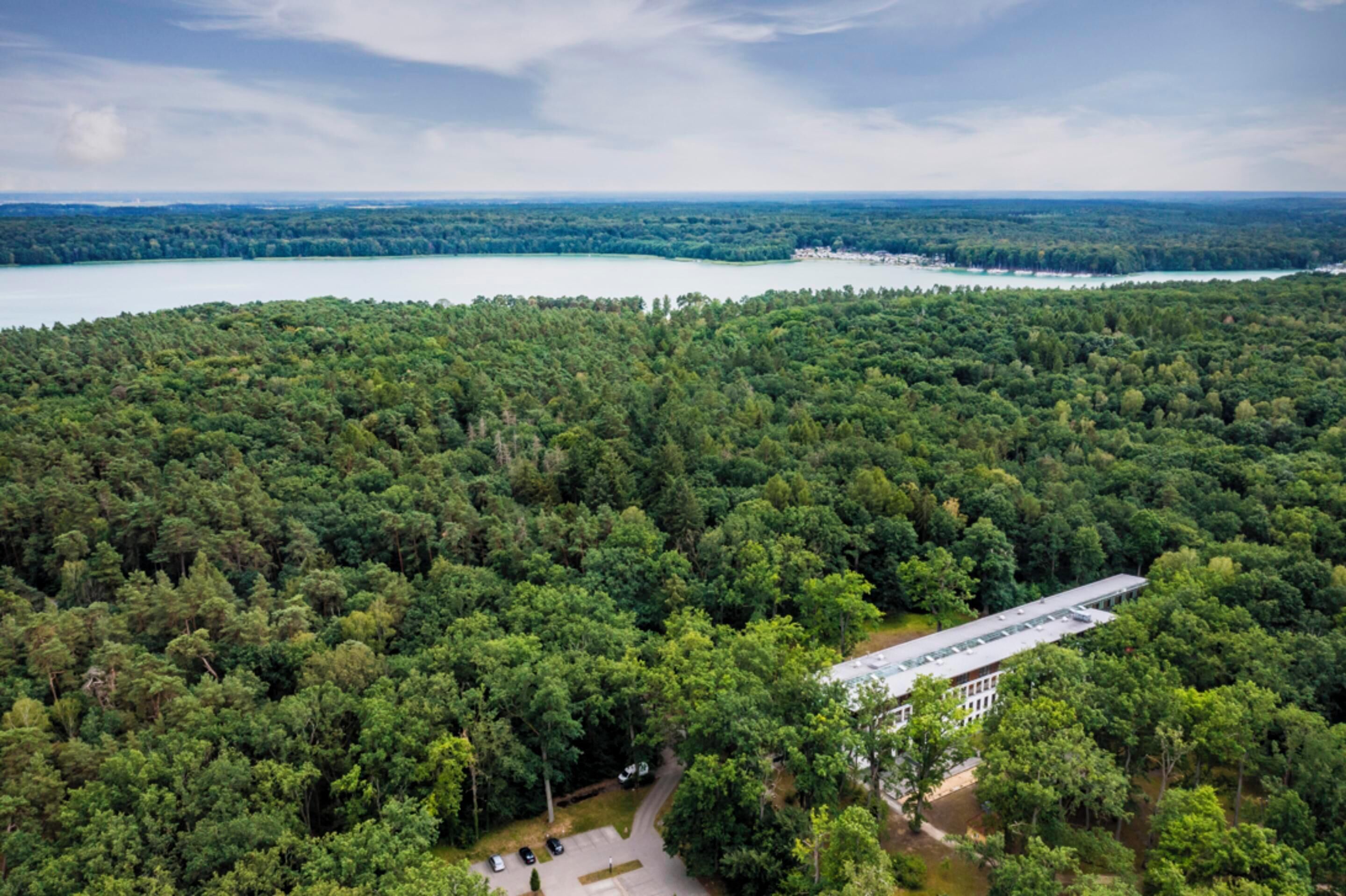 An aerial view of a forested area with a building that appears to be a lodge or resort, surrounded by dense trees and a lake in the background.