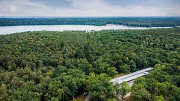 An aerial view of a forested area with a building that appears to be a lodge or resort, surrounded by dense trees and a lake in the background.