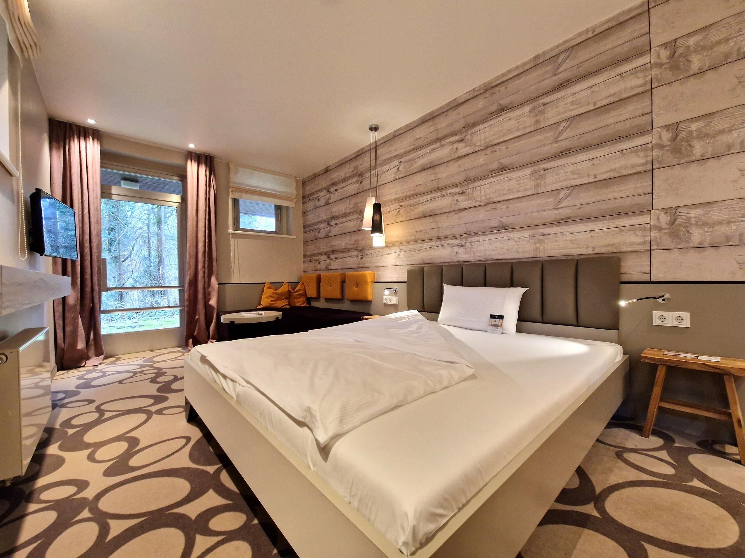Una habitación de hotel moderna con una cama grande, un panel de madera decorativo, un escritorio pequeño y una ventana con cortinas.