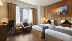 Ein Hotelzimmer mit zwei Betten, einem Schreibtisch, einem Stuhl und einem fensterbepflanzten Fenster.