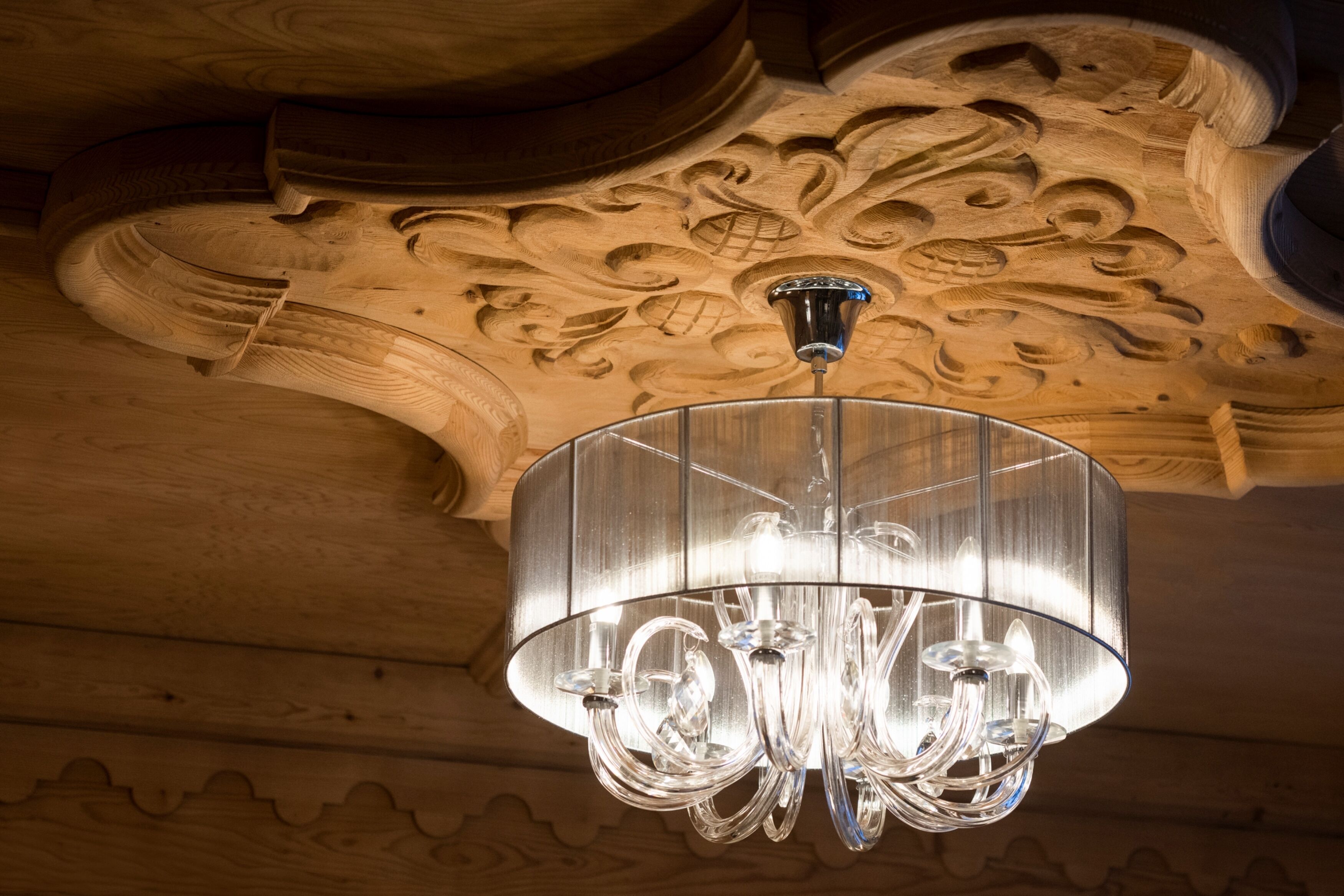 Um candelabro com cúpula de vidro transparente pendurado em um teto de madeira com esculturas detalhadas.