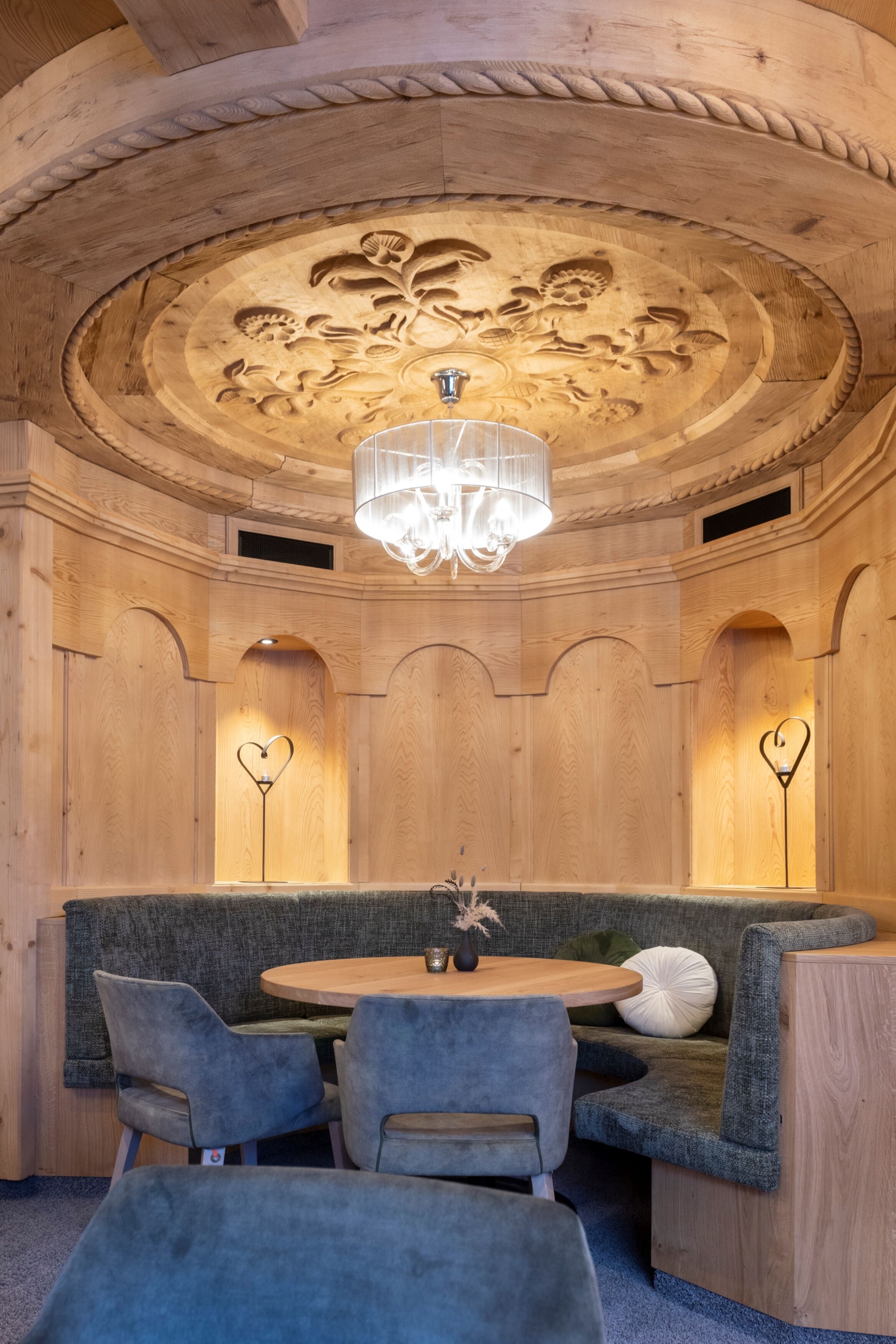 A wooden interior with a round table, grey upholstered seating, and a chandelier.