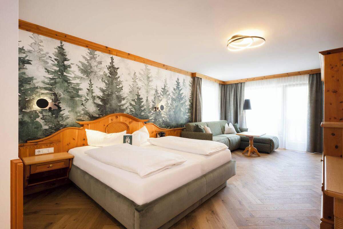 Une chambre d’hôtel avec un grand lit, une tête de lit en bois, une table de chevet, un canapé vert, une petite table et une fresque murale représentant des arbres.
