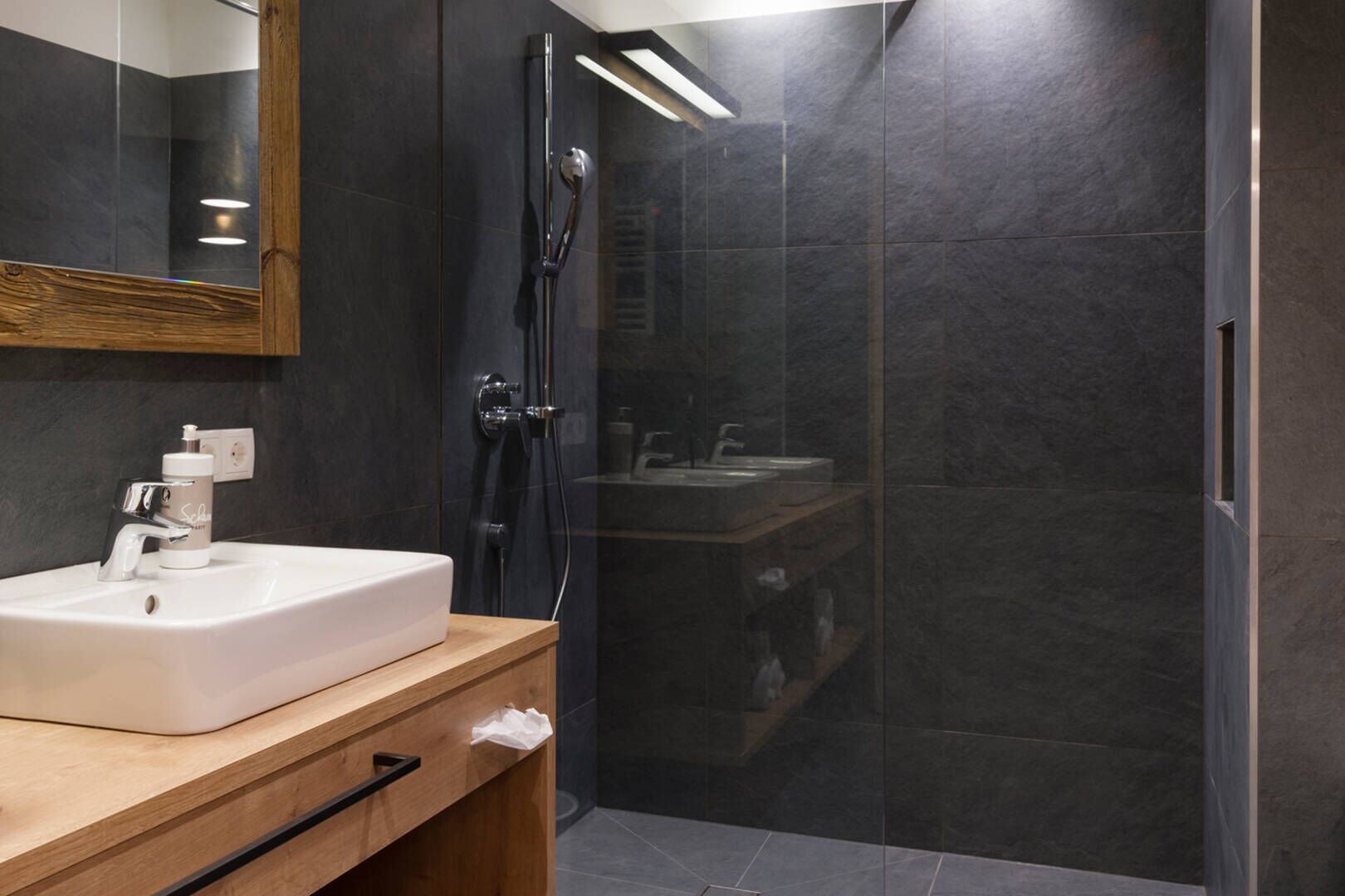Un baño moderno con un lavamanos blanco, baldosas oscuras y una cabina de ducha de cristal.