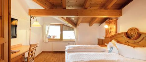 A bedroom with wooden beams, a bed, a desk, and a window.