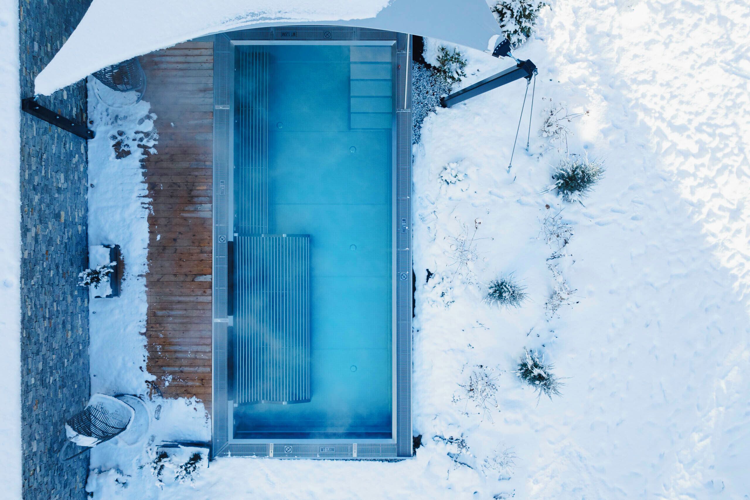 A rectangular swimming pool with a wooden deck and metal railings, surrounded by snow.
