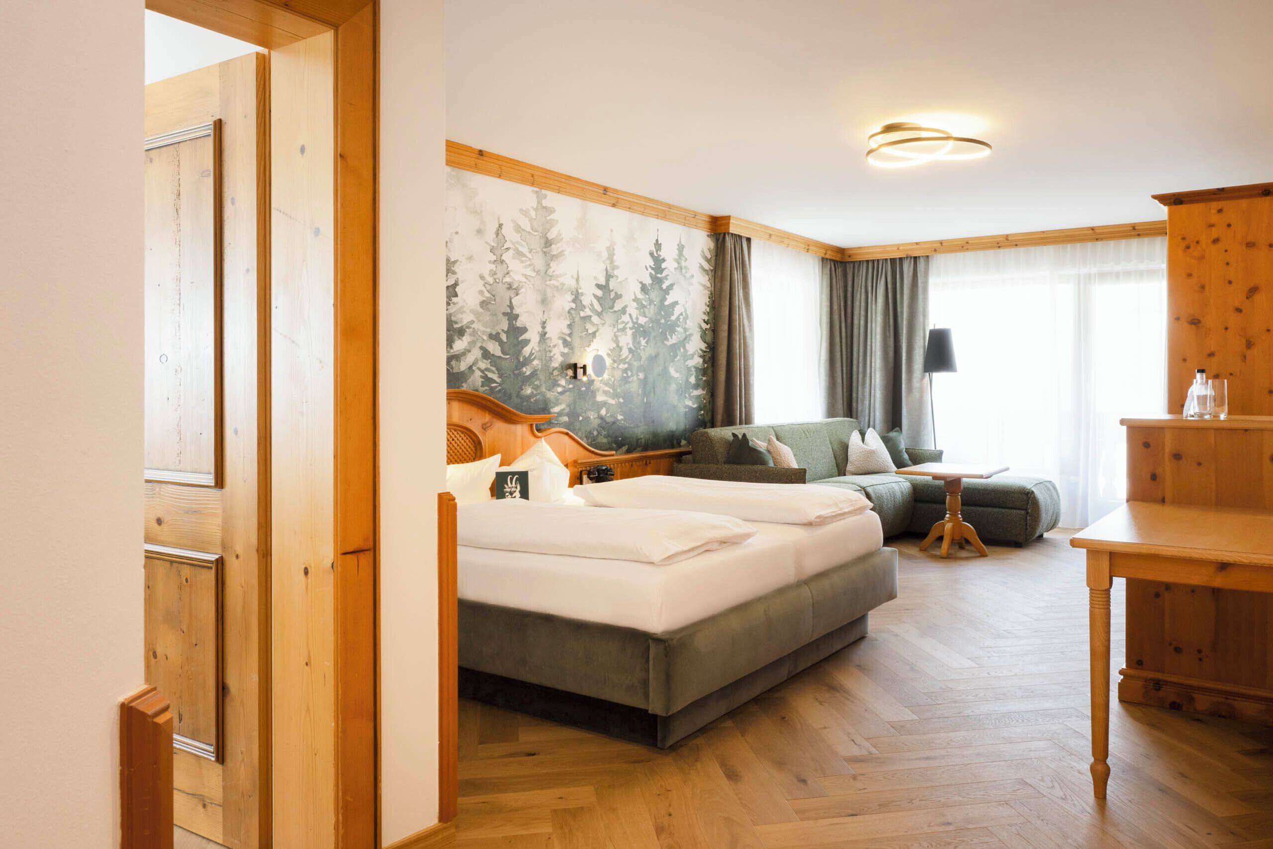 A hotel room with a bed, a sofa, a wooden table, and a wall with a forest mural.
