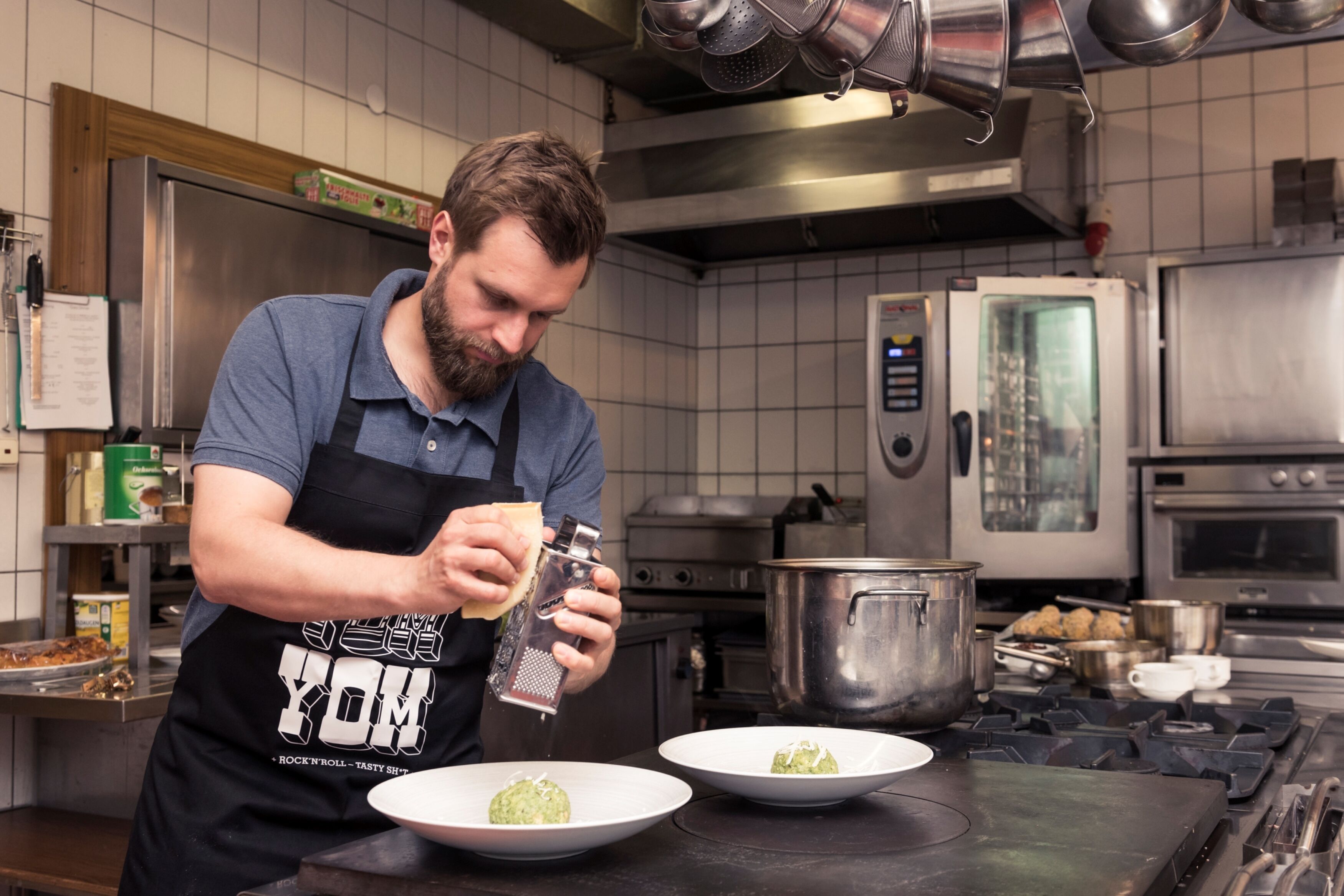 A chef grating cheese over a dish in a kitchen with stainless steel appliances and tiled walls.