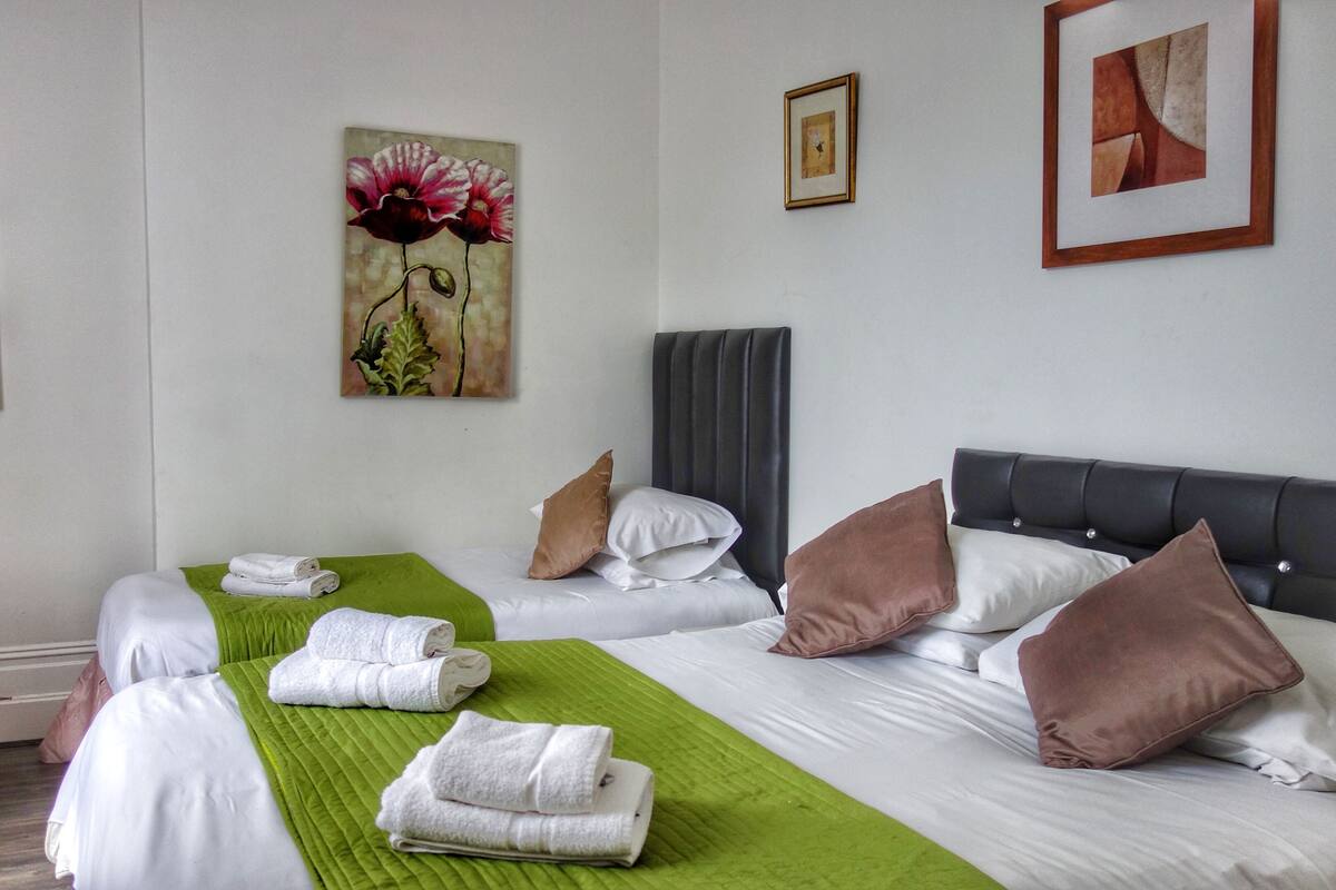 A hotel room with a bed, green bedspread, white linens, brown pillows, and two paintings on the walls.