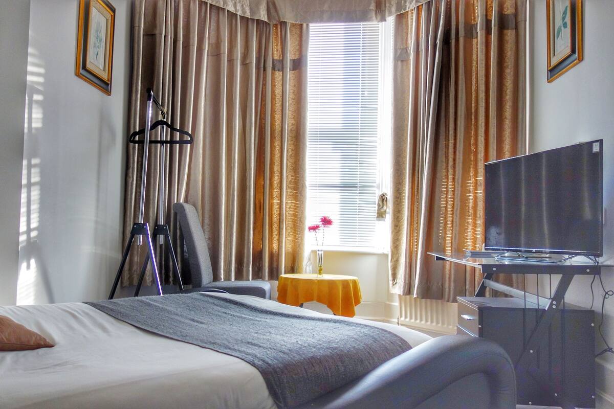 A hotel room with a bed, a desk with a computer, a window with curtains, and a television.