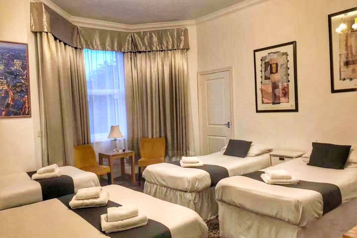 A hotel room with two single beds, a small table, a chair, and a window with curtains.