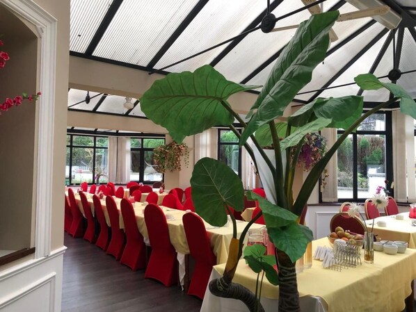 A restaurant with a long table set for a meal, large green plants, and red chairs.