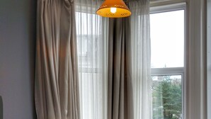 A neatly made bed with pillows and a blanket, a bedside lamp, and a hanging lamp above it. There are curtains on the window, and a view of the outside is visible. - The Kensington Hotel (Bournemouth)